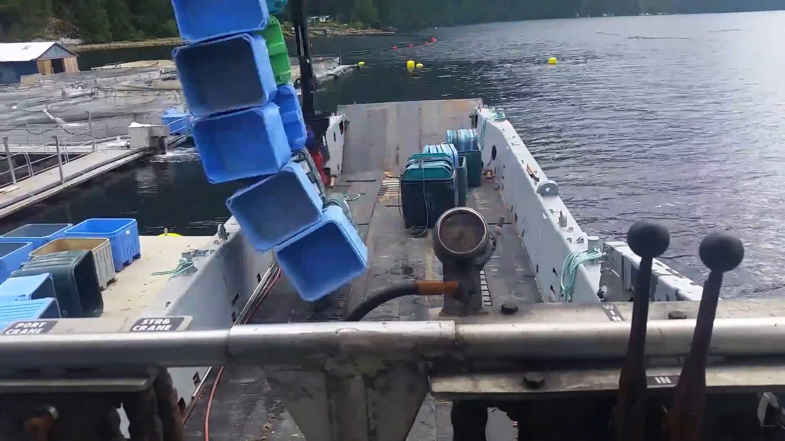 Looking forward over the Inlet Raider's deck: a stack of blue totes hangs from the crane beside a float with more totes on it, calm water and a forested shore behind