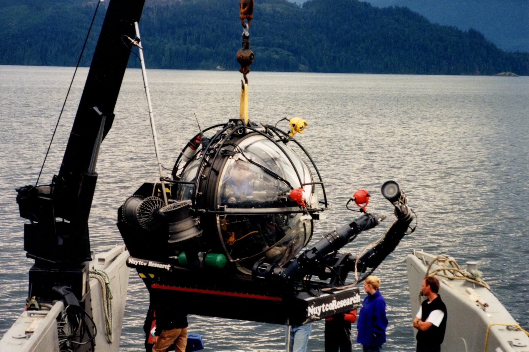 A research submersible with a clear sphere hangs from a crane hook over a landing craft's deck, people watching from the deck, a forested shore across the water