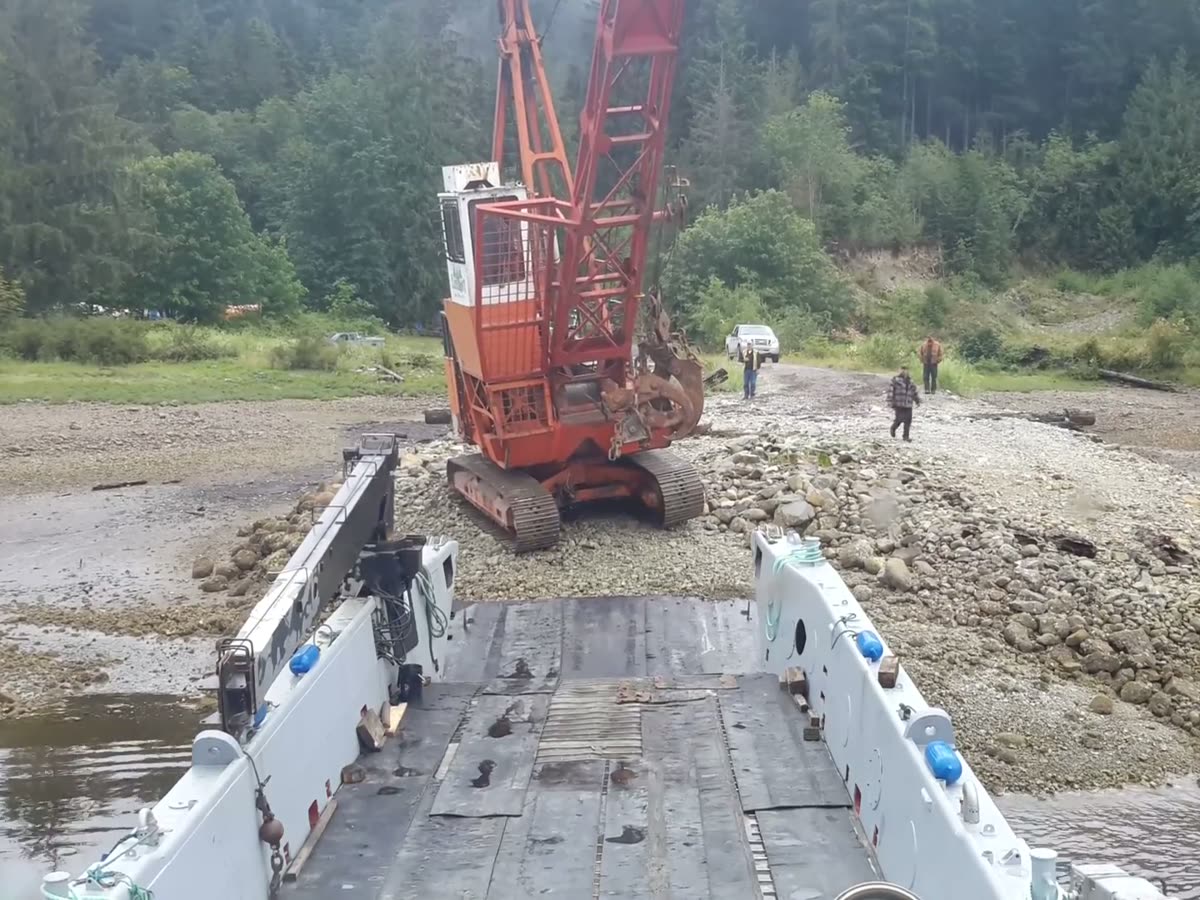 An orange grapple yarder on a gravel beach, lined up with the lowered bow ramp
