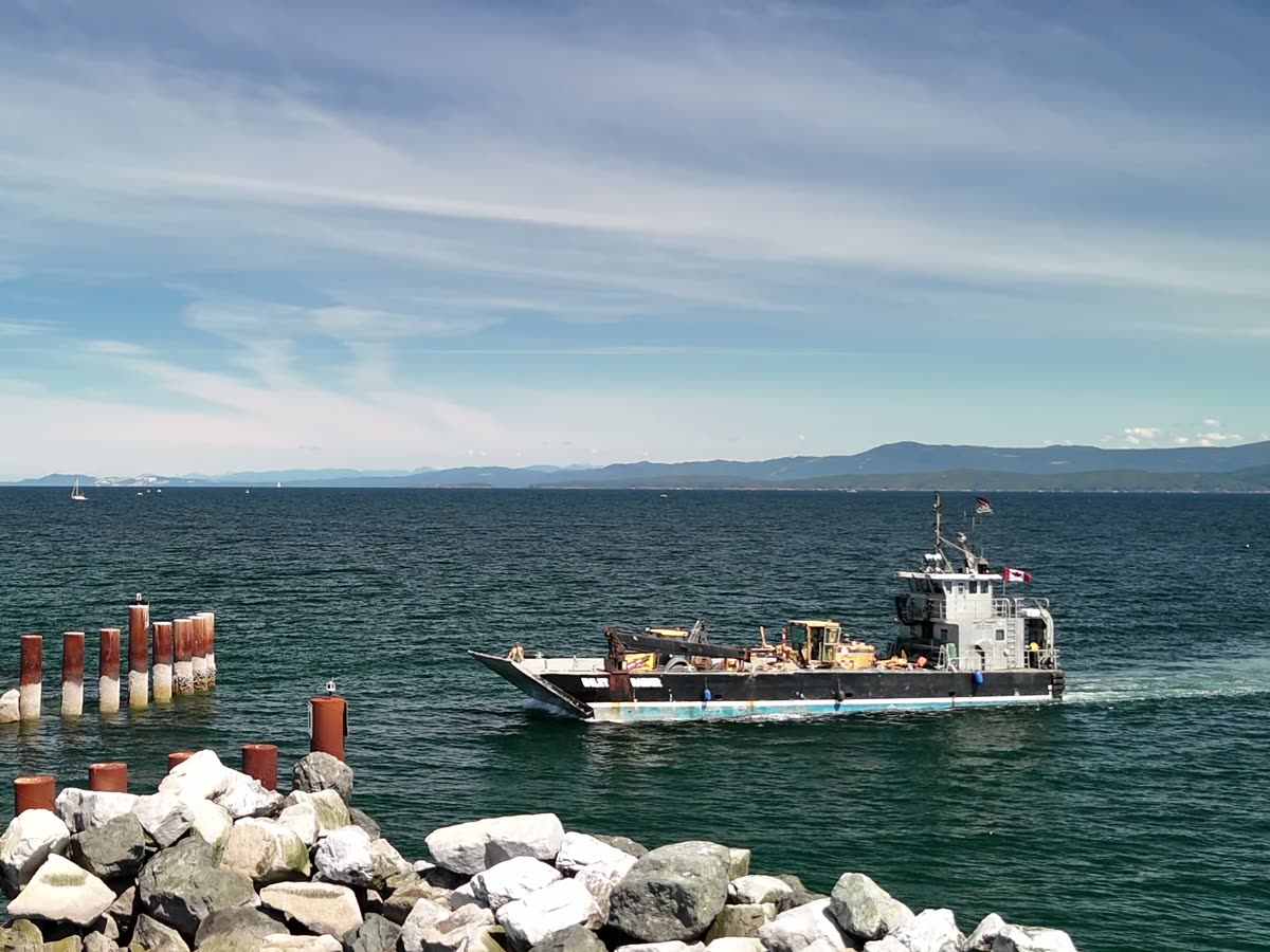 The Inlet Raider with a machine on deck, passing a rock breakwater and pilings