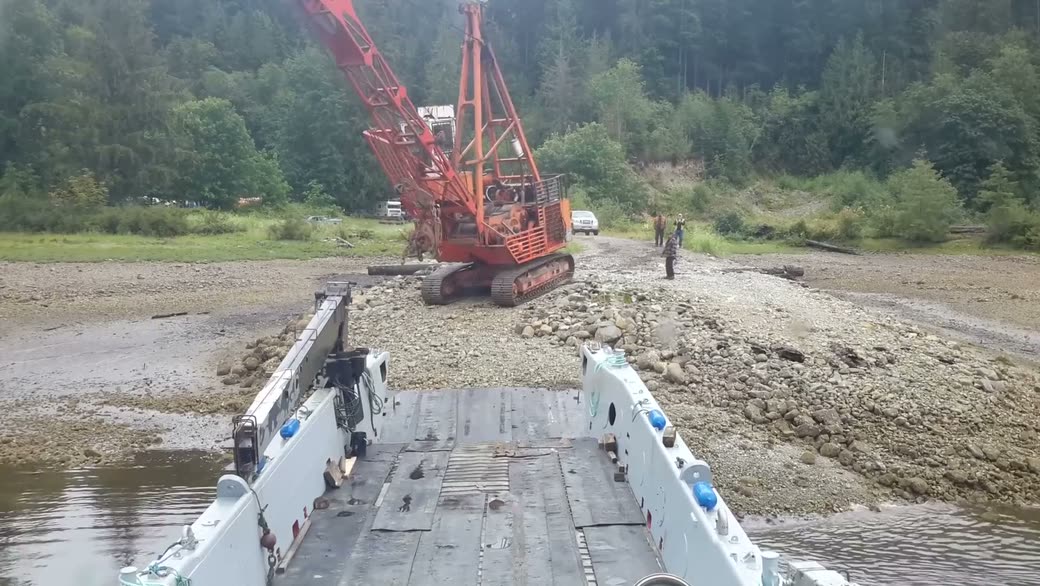 An orange grapple yarder on a gravel landing, seen over the lowered bow ramp